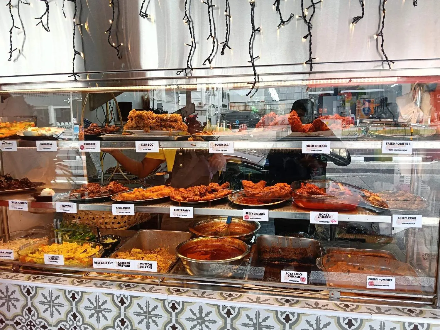 The Yaseem Nasi Kandar counter on MacPherson Road in daylight — steel trays of curries, fried chicken and pomfret behind glass, each under its printed bilingual label